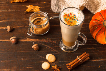 Latte in a tall spiced glass on a dark wooden table with dried maple leaves, pumpkin and cinnamon sticks and cookies