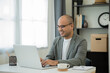 © Chanakon - A middle-aged man around the age of 35. Working at home Work through the laptop by meeting video conference. He was wearing a grey suit and glasses. Smiling asian businessman work from home.