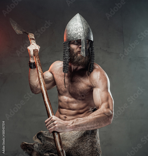 Dangerous and naked nordic barbarian with beard and helmet on him posing in dark background with two handed axe.