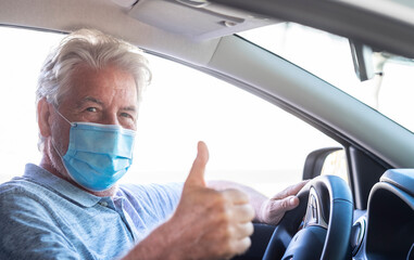 A senior man with white hair wearing surgical mask gesturing ok with hand while driving the car