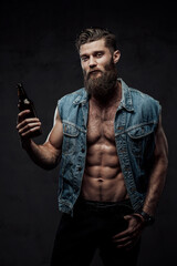 Dressed in modern casual clothing bearded and muscular guy with stylish haircut posing holding beer bottle in dark background.