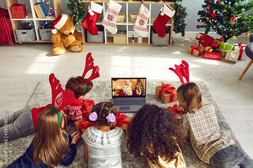 A group of children lying on the floor watching a video chat with Santa using a laptop in a decorated room during Christmas and New Years.