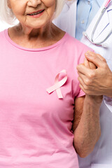 Wall Mural - Cropped view of doctor and senior woman with pink ribbon on t-shirt holding hands isolated on white