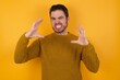 © Roquillo - Young man wearing casual sweater and over isolated yellow background Shouting frustrated with rage, hands trying to strangle, yelling mad.