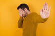 © Roquillo - Young man wearing casual sweater and over isolated yellow background covers eyes with palm and doing stop gesture, tries to hide from everybody.