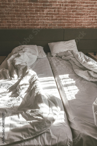 Unmade two beds with white blankets and pillows. Loft apartment with red brick wall. Minimalist or scandinavian style of interior design. Grunge look of house. Spacious bedroom with furniture