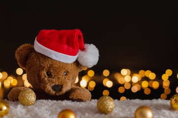 brown teddy bear wearing santa claus hat with christmas ornaments and lights background.
