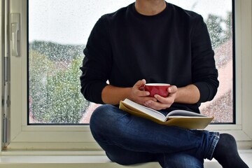 A man in his mid-twenties sits on a windowsill, in the background raindrops can be seen on the window pane - the man is reading a book while drinking a coffee - anonymous picture without head and face