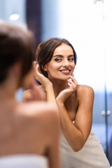 Wall Mural - Smiling young woman in bathrobe and towel on head looking at mirror in bathroom