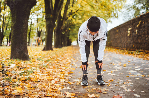 Mid aged fit athletic woman dressed modern running clothes standing and bend over to stretch the back on the footway after jogging in the autumnal city park. Active running people concept image.