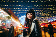 © bodnarphoto - Pretty smiling girl stands on a Christmas decorated street in evening with a hot drink in her hand and poses for camera with a smile on face. Girl at the evening fair with lanterns on background.