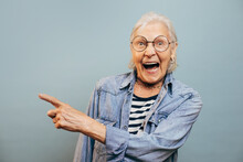 Emotional Very Excited Old Woman Indexing At Something By Her Finger And Laugting. Cool Stylish Senior Woman Looking At Camera, Wearing Denim Jacket, Striped Shirt. Isolated Over Blue Backgound.