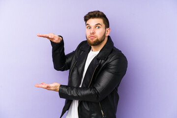 Young man isolated on purple background shocked and amazed holding a copy space between hands.