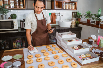 Smiling chef male wearing an apron using a smartphone standing while write to note front donuts on a table in the kitchen