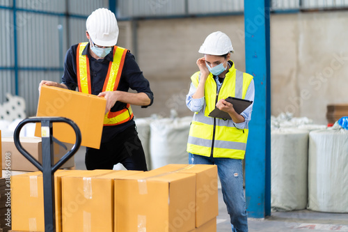 Female Inventory Manager checking stock on Digital Tablet. Man warehouse worker loading or unloading boxes with hard hat safety helmet at face mask factory.