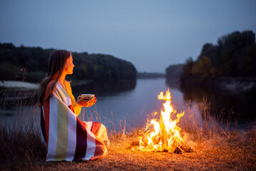 Beautiful girl admiring the beauty of nature. Lonely dreamy girl drinking hot tea, sitting near bonfire on the edge of the river