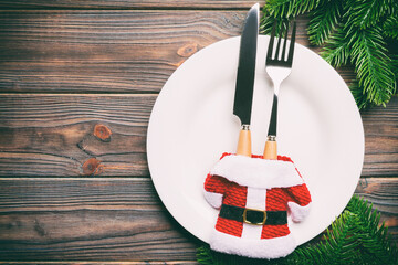 Sticker - Holiday composition of plate and flatware decorated with Santa clothes on wooden background. Top view of Christmas decorations with empty space for your design. Festive time concept