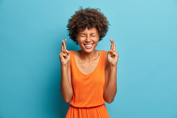 Wall Mural - Superstitious cheerful African American woman keeps fingers crossed wishes for good luck believes in dreams come true laughs with closed eyes dressed in orange summer dress isolated on blue background