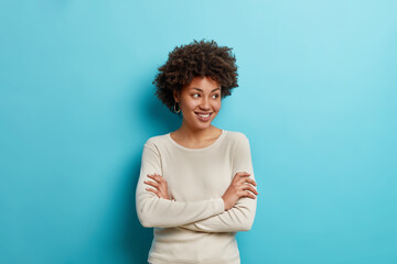 Poster - Smiling relaxed dark skinned woman stands with crossed arms and looks away happily dressed in casual clothes enjoys happy moment stands in thoughtful pose isolated over blue studio background.