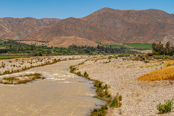 Vicuna, Chile - December 7, 2008: landscape with brown water fast streaming Elqui River with band of green agriculture and trees separating plain from mountain range under blue sky.