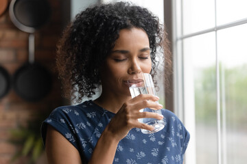 Wall Mural - Happy young African American woman feel dehydrated thirsty drink clean pure mineral water from glass at home. Millennial biracial female enjoy clear aqua for body refreshment. Hydration concept.