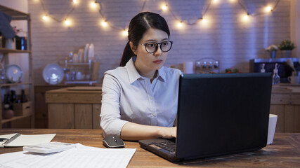 Wall Mural - Young asian korean businesswoman working on laptop computer at late night home kitchen. concentrated female typing on keyboard of notebook pc with document paper and calculator beside on wood table