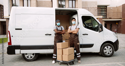 Portrait of African American and caucasian delivery men in mask and uniform in front of his van.