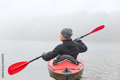 Fotomural Back view of young handsome man kayaking in river, spends foggy morning in canoe, extreme sport, man in black jacket and gray cap rowing boat and looking aside