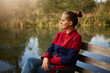 Side view of pensive woman sitting on wooden bench on bank of river, lady with knot looking at water and dreams, wearing casual attire, enjoys beautiful nature.