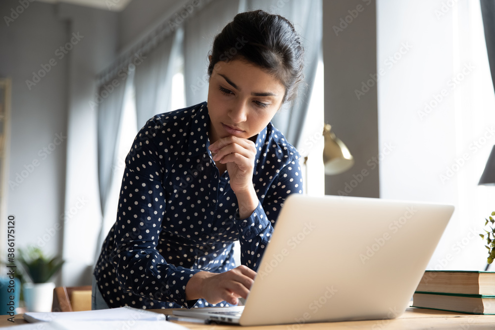Pensive indian business woman standing in office room standing lean over workplace desk working on laptop thinking about problem solution, create research, do paperwork, corporate task prepare report - obrazy, fototapety, plakaty 