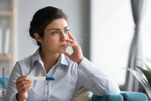 Indian ethnicity woman holding pregnancy test looking out the window deep in sad thoughts about health problems and infertility feels troubled by result, bad news, unwanted pregnancy, abortion concept