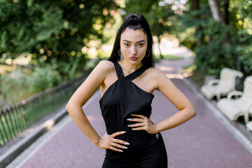 Outdoor portrait of attractive young Asian mixed raced girl with black ponytail hair, wearing elegant fashionable black clothes, posing to camera, posing in the green park alley