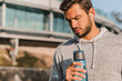 © InsideCreativeHouse - Sporty handsome sportsman with beard and earbuds holding bottle of water behind urban landscape