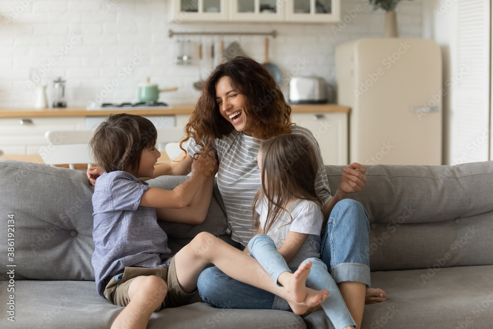 Overjoyed young mother having fun with two kids at home, laughing happy mum, adorable little daughter and son playing funny active game, tickling, sitting on cozy couch in living room - obrazy, fototapety, plakaty 