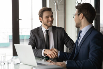 Canvas Print - Smiling two young businessman in formal suits shaking hands, making agreement after online project presentation on computer. Happy manager thanking employee for presenting development strategy.