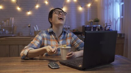 Wall Mural - relax young asian japanese girl eating instant noodles and laughing cheerfully while watching online funny video on laptop computer