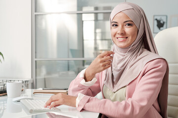 Poster - Cheerful Muslim businesswoman in earphones listening to online course by desk