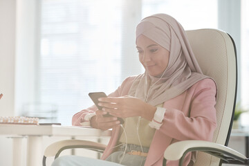 Poster - Young smiling Muslim businesswoman in hijab scrolling in smartphone in office
