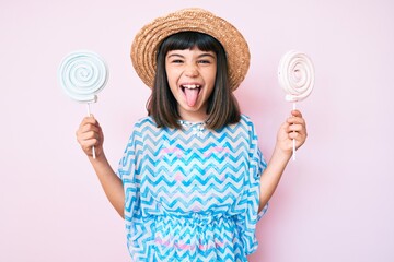 Young little girl with bang wearing summer dress eating candy sticking tongue out happy with funny expression.