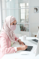 Poster - Young creative female designer in hijab and protective mask sitting by computer