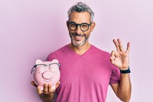 Middle Age Grey-haired Man Holding Piggy Bank With Glasses Doing Ok Sign With Fingers, Smiling Friendly Gesturing Excellent Symbol