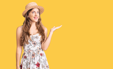 Wall Mural - Beautiful caucasian young woman wearing summer hat smiling cheerful presenting and pointing with palm of hand looking at the camera.