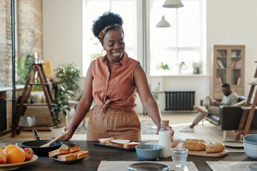 Canvas Print - Young smiling African woman in casualwear taking bottle of milk while cooking