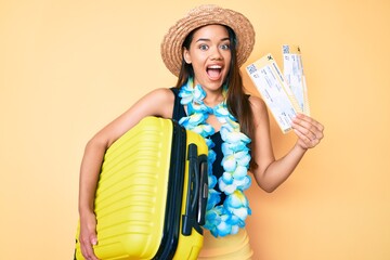 Young beautiful latin girl wearing summer hat and hawaiian lei holding cabin bag and boarding pass celebrating crazy and amazed for success with open eyes screaming excited.