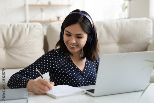 Happy young Asian woman in headphone write make notes study distant on computer from home. Smiling Vietnamese female in earphones take online course or training on laptop, watch webinar on gadget.