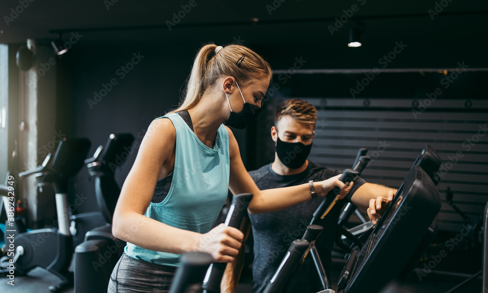 Young fit and attractive woman at body workout in modern gym together with her personal fitness instructor or coach. They wearing protective face masks. Coronavirus world pandemic and sport theme. - obrazy, fototapety, plakaty 