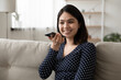 © fizkes - Smiling young Asian girl sit on sofa at home record audio message on smartphone talking online. Happy Vietnamese woman speak chat on cellphone using loudspeaker, activate voice recognition app.