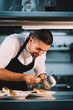 © Henko Studio - Portrait of a male chef decorating food with spoon in ceramic dish over stainless steel worktop in restaurant kitchen.