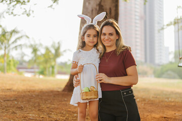 Wall Mural - Latin family celebrating Easter. curly girl with bunny ears and holding a basket of easter eggs hugging her mother in the park