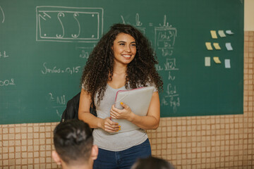 Latin Curly haired student smiling wearing backpack holding a notebook in a classroom